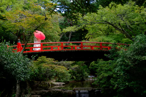 A person in white holding a red parasol stands on a bright red arched bridge surrounded by lush green trees over a calm stream in a peaceful garden setting.