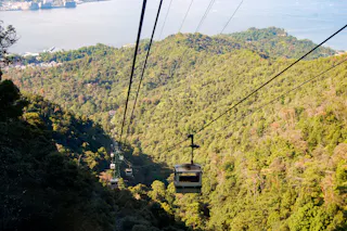 A cable car travels above a lush, green forested mountain slope with other cable cars visible behind it; a body of water and coastline appear in the background.