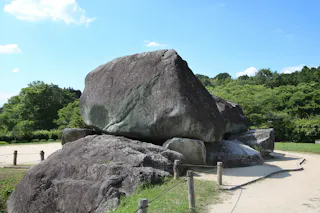 A large, ancient stone structure made of several stacked rocks, surrounded by a sandy path and greenery under a clear blue sky.