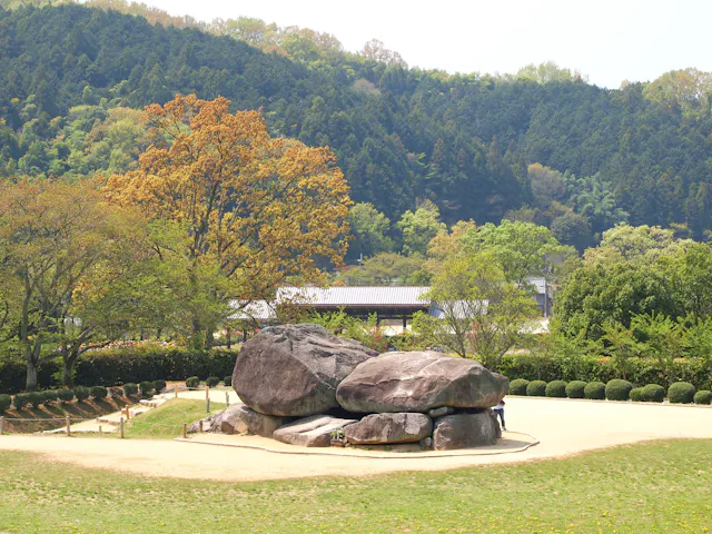 Large ancient rocks form a low stone structure in a grassy area, surrounded by trees and dense forested hills in the background, under a bright sky.