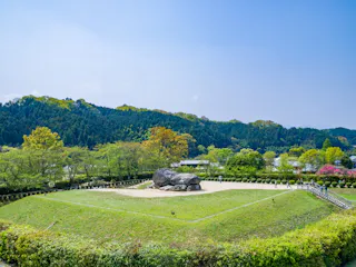 A large ancient stone structure sits on a grassy, raised mound surrounded by trimmed bushes and trees, with a backdrop of forested hills under a clear blue sky.