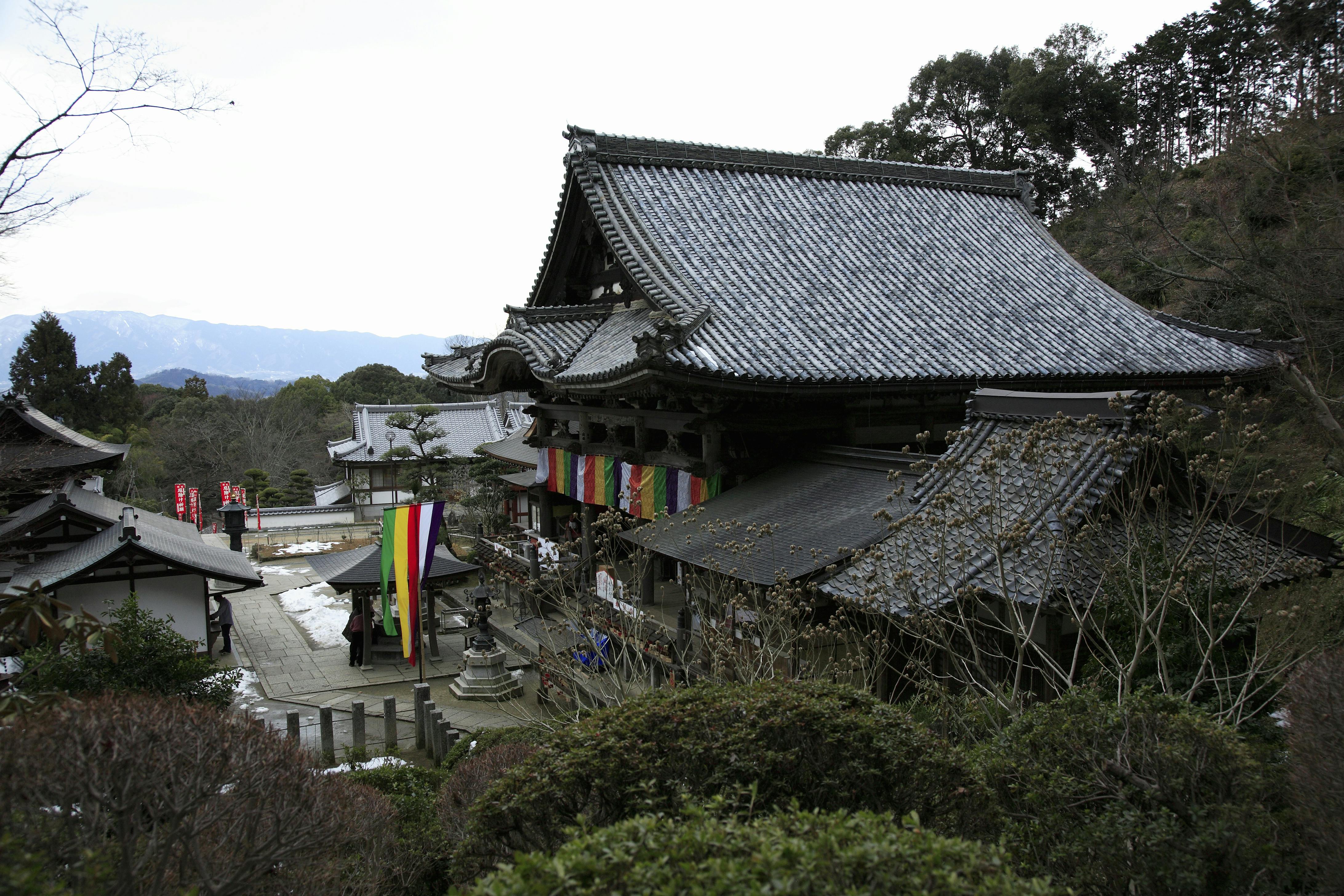 Traditional Japanese temple buildings with tiled roofs and colorful banners hang on the main structure, surrounded by trees and shrubs in a hilly landscape under a cloudy sky. Visitors are visible near the entrance.