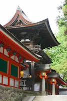 A traditional Japanese shrine with ornate wooden roofs, red pillars, green panels, and stone steps. Lanterns line the pathway, and lush greenery surrounds the building.
