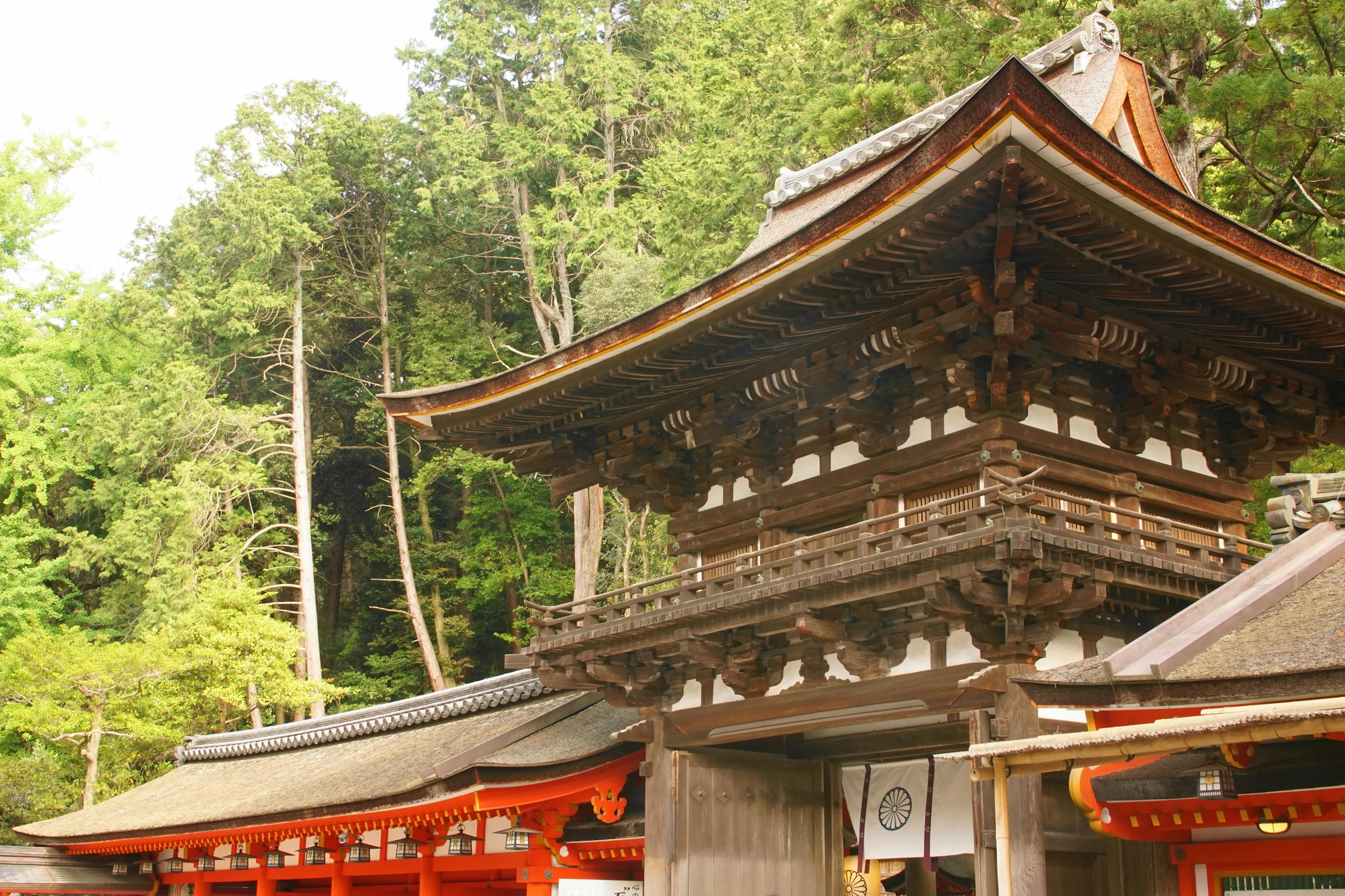A traditional Japanese temple with ornate wooden architecture stands surrounded by tall green trees. The roof features layered eaves and decorative details, while red and white structures frame the entrance below.