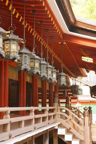 A row of ornate metal lanterns hangs from the eaves of a traditional Japanese shrine with red beams, wooden railings, and green accents.