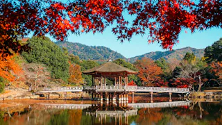 A traditional Japanese pavilion stands over a calm pond, surrounded by vibrant autumn trees and mountains. Red maple leaves frame the top of the image under a clear blue sky.