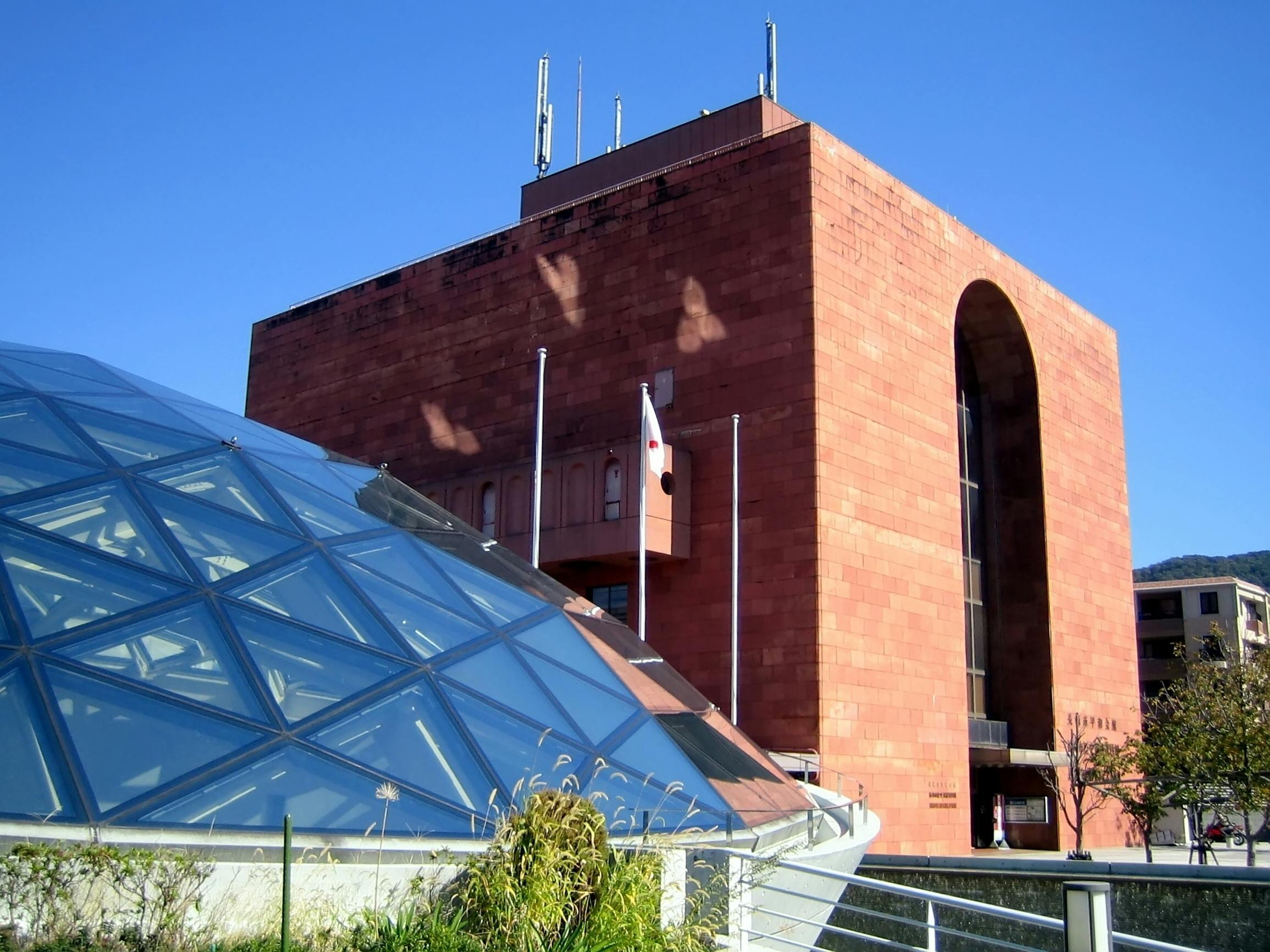 A modern glass dome structure sits in front of a large red-brick building with a distinctive arched window, under a clear blue sky.