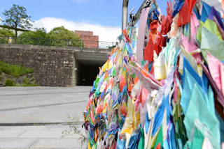 Colorful paper cranes hang in clusters in the foreground, symbolizing peace. In the background, there is a stone wall, greenery, and a partially cloudy sky over an outdoor urban area.