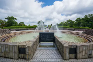 A circular stone fountain with jets of water, surrounded by greenery and trees under a partly cloudy sky. There are plaques with inscriptions and people walking in the background.