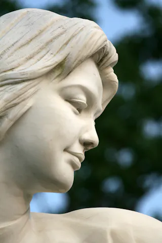 Close-up of a white marble statue depicting a serene woman's face, shown in profile against a blurred background of green foliage and blue sky.