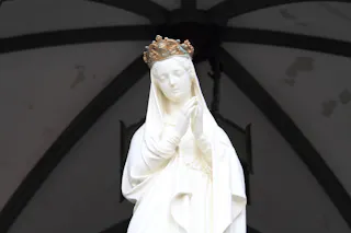 A white statue of a woman wearing a crown, with her hands clasped in prayer and eyes closed, stands against a background of arched beams and a lightly stained ceiling.