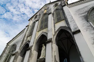 Low-angle view of a large church facade with tall arched windows, weathered white walls, decorative details, and a statue of a religious figure at the entrance under a partly cloudy sky.