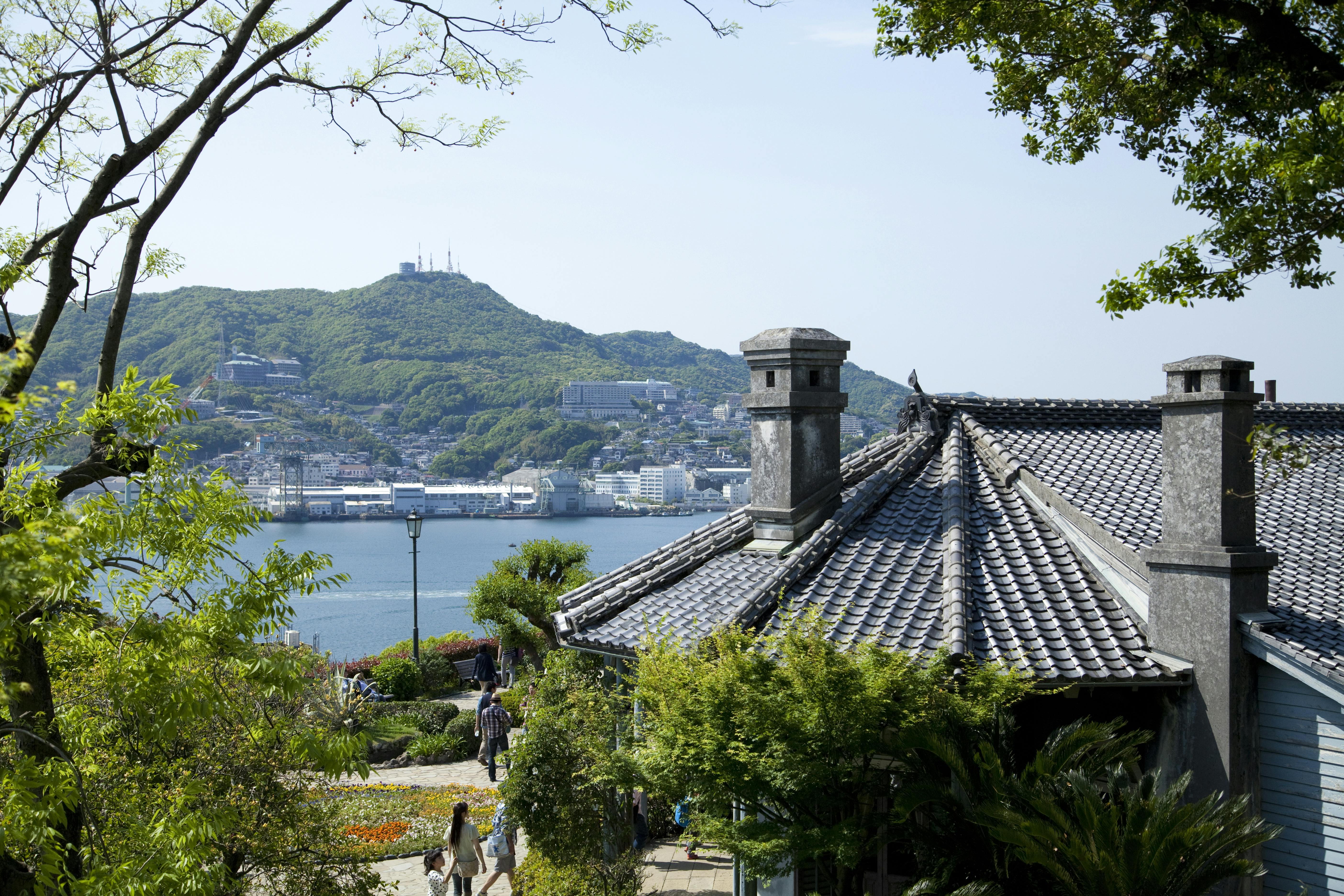 A traditional Japanese building with a tiled roof is surrounded by greenery, overlooking a waterfront with mountains and a town in the background under a clear sky.