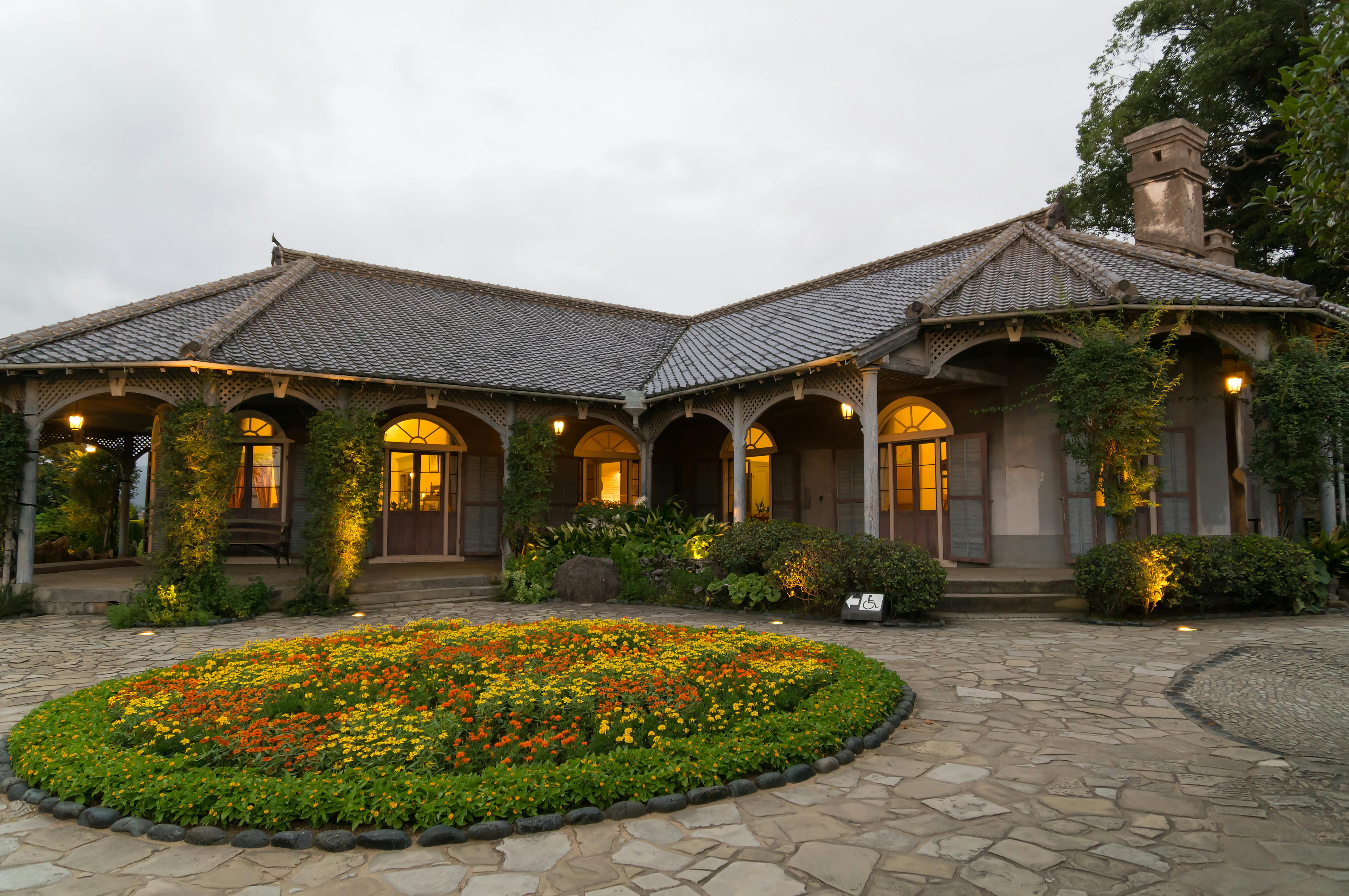 Historic single-story building with a tiled roof, arched windows, and warm yellow lights, surrounded by lush greenery and a round flower bed with colorful blooms on a stone-paved courtyard at dusk.
