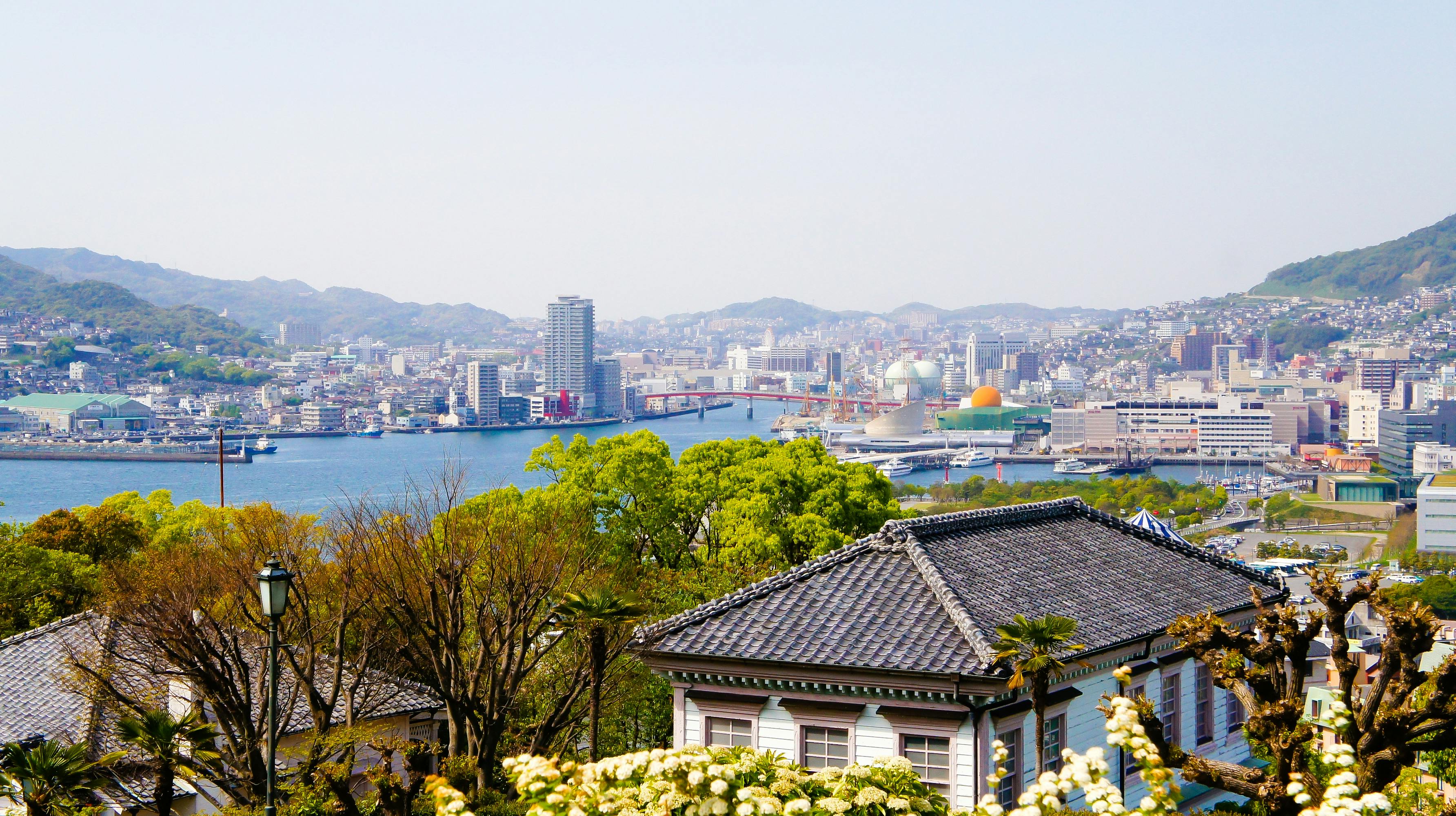 Panoramic view of a coastal city with mountains in the background, a river running through, modern and traditional buildings, greenery, and a clear sky.