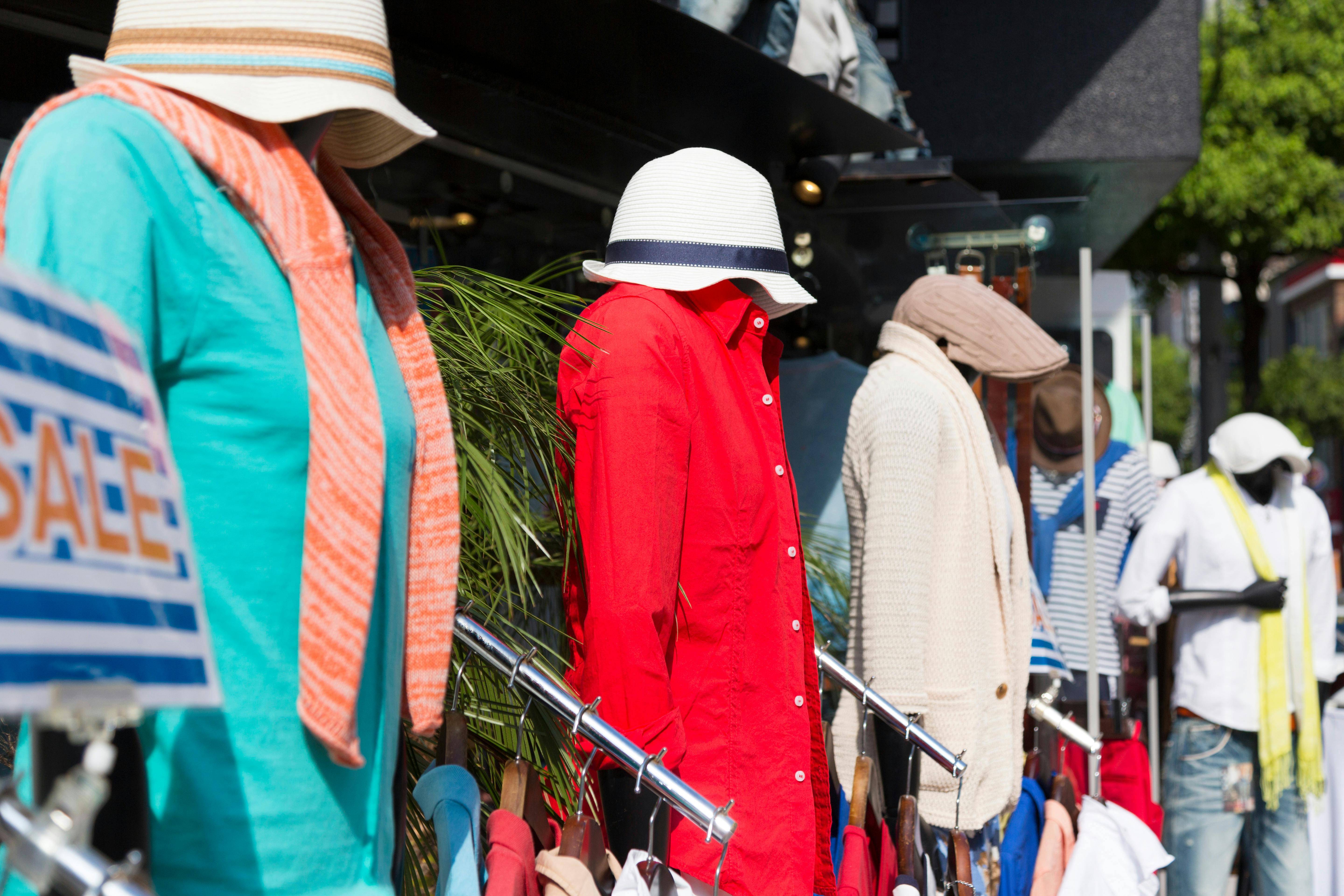 Mannequins dressed in colorful casual clothes and hats are displayed outside a store in bright sunlight, with a "SALE" sign visible on the left side. Trees and buildings appear in the background.