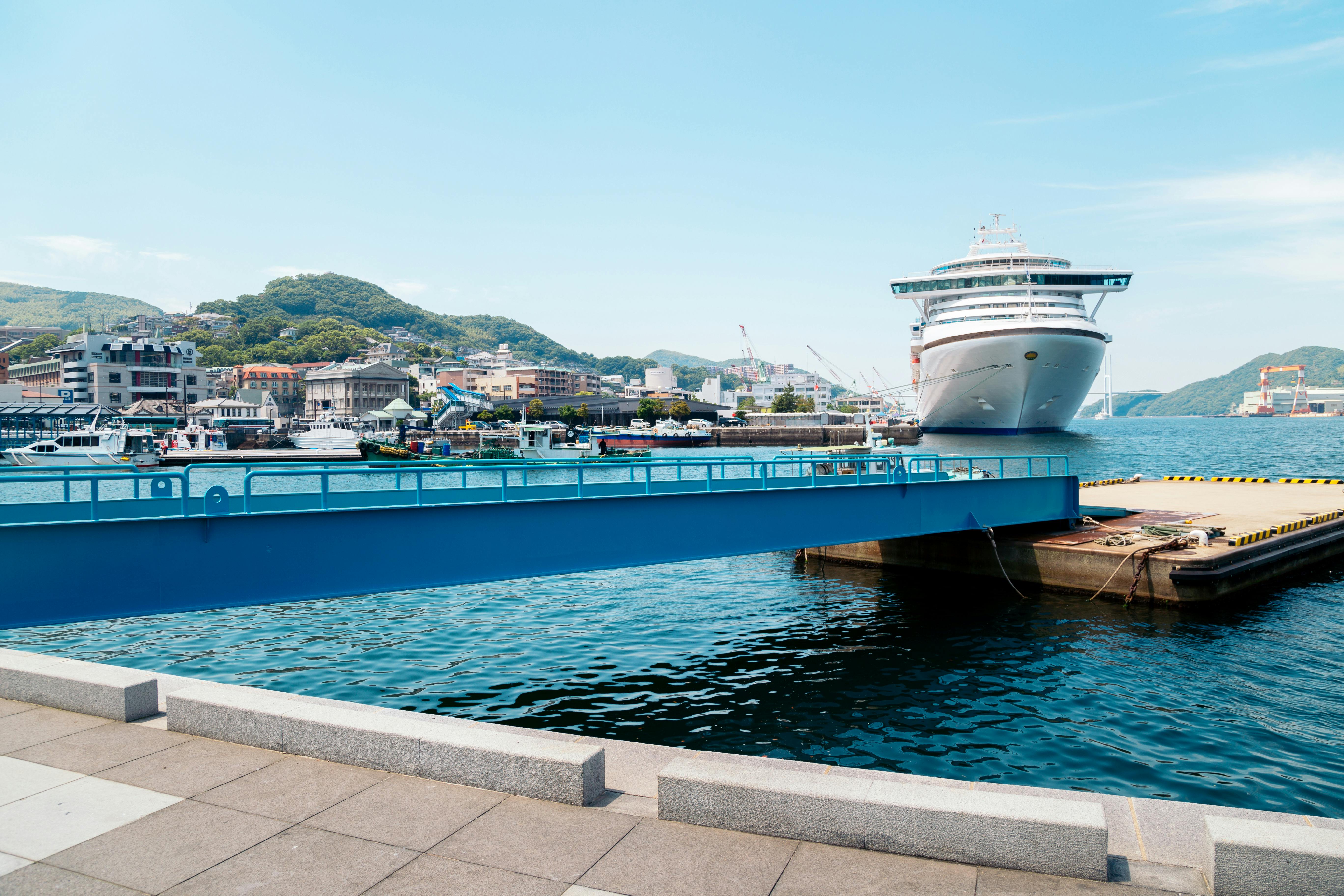 A large white cruise ship is docked at a port near a coastal town with hills in the background. The scene includes a blue pedestrian bridge over the water and a clear, sunny sky.