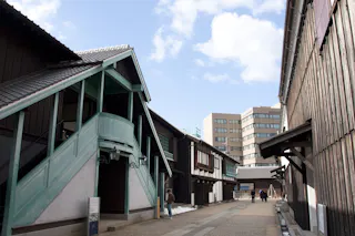 A pedestrian walkway lined with traditional Japanese wooden buildings on the left and right, with modern buildings visible in the background under a partly cloudy sky. A few people are walking along the path.