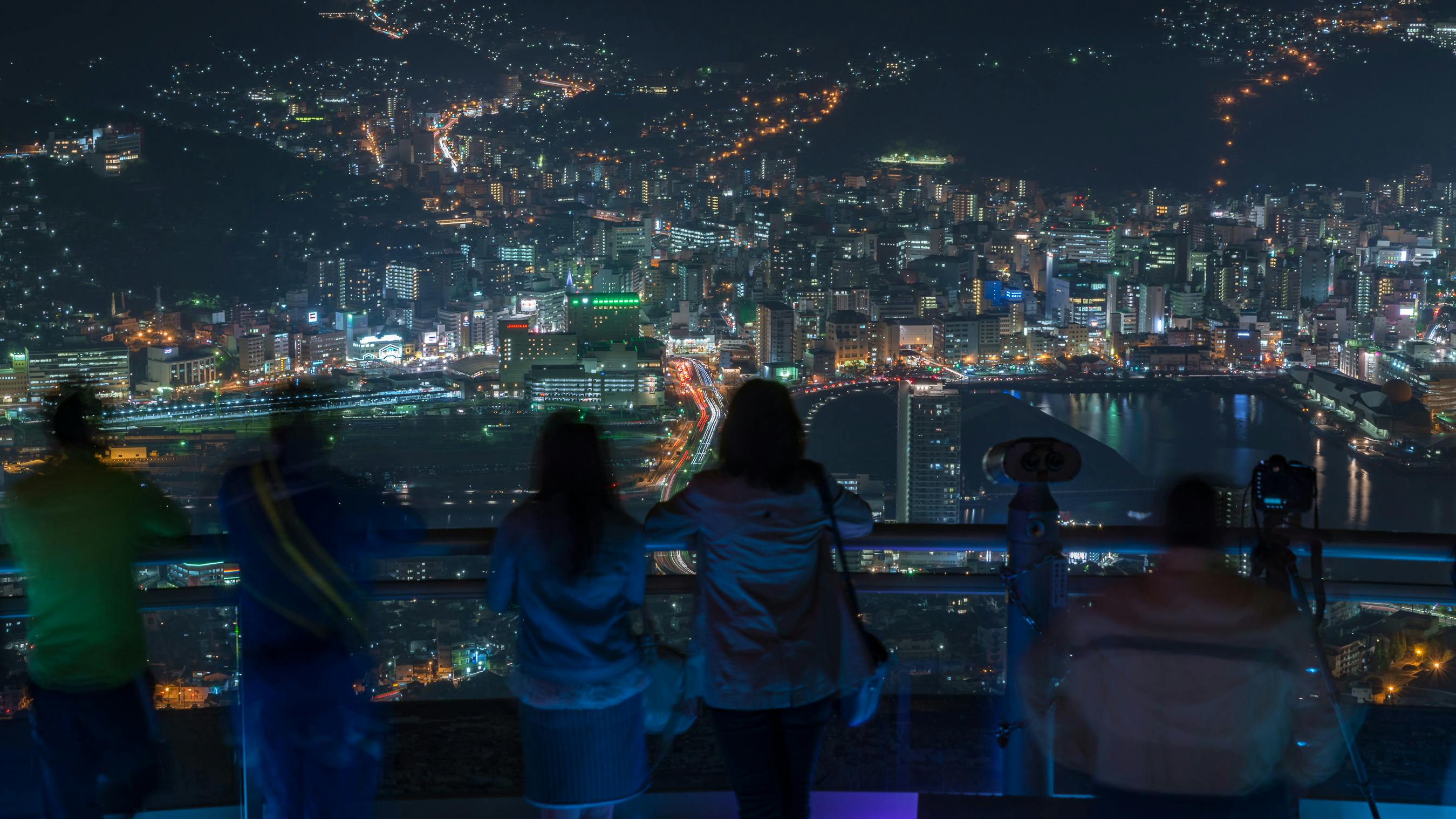 Several people stand at an observation deck at night, looking out over a brightly lit cityscape filled with buildings, winding roads, and clusters of lights stretching into the distance.