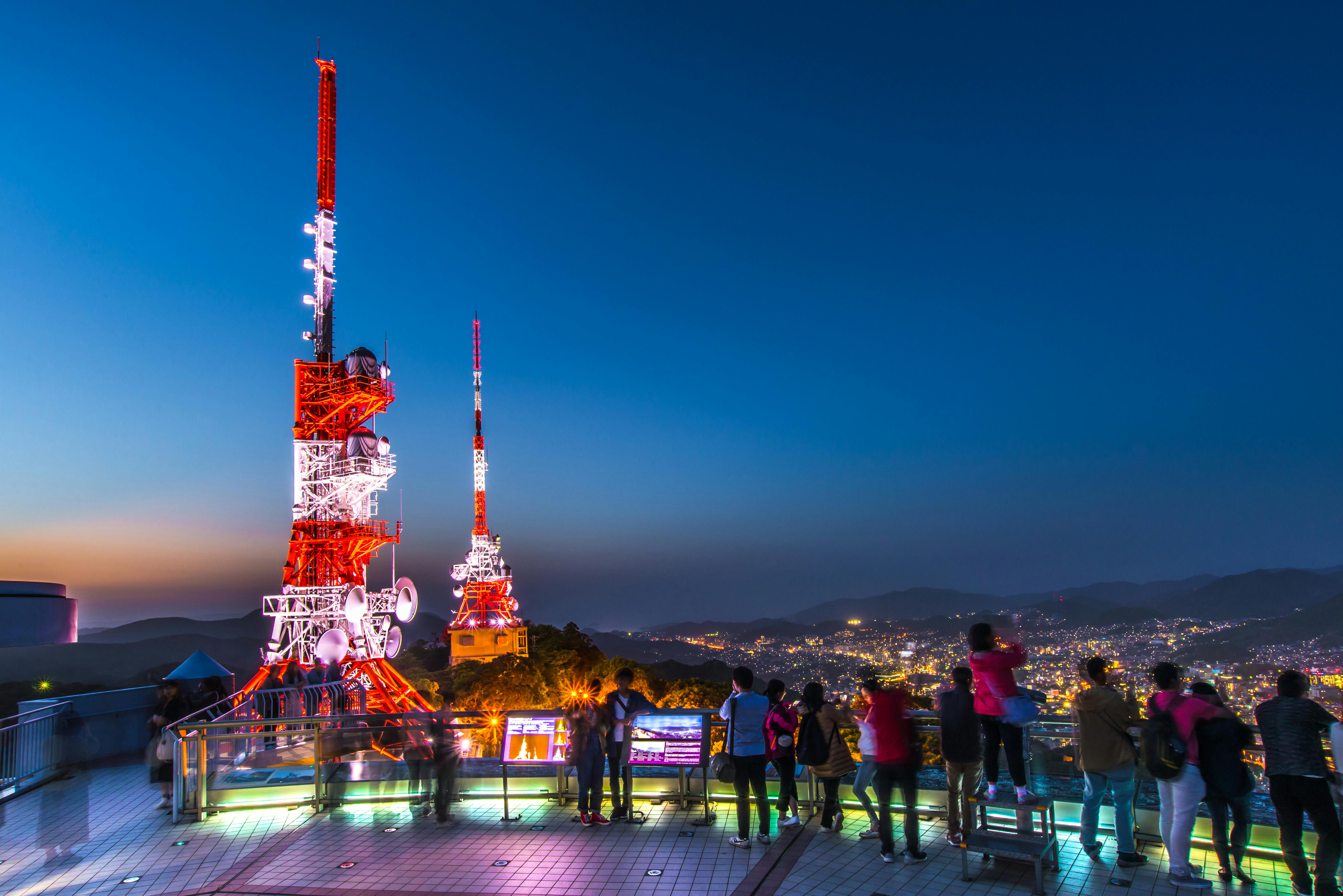 People stand on an observation deck at night, viewing a brightly lit red-and-white broadcast tower and a cityscape illuminated below, with mountains visible in the distance under a clear sky.