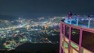 People stand on a brightly lit observation deck at night, overlooking a cityscape filled with glowing buildings, roads, and water, all surrounded by dark hills and mountains.