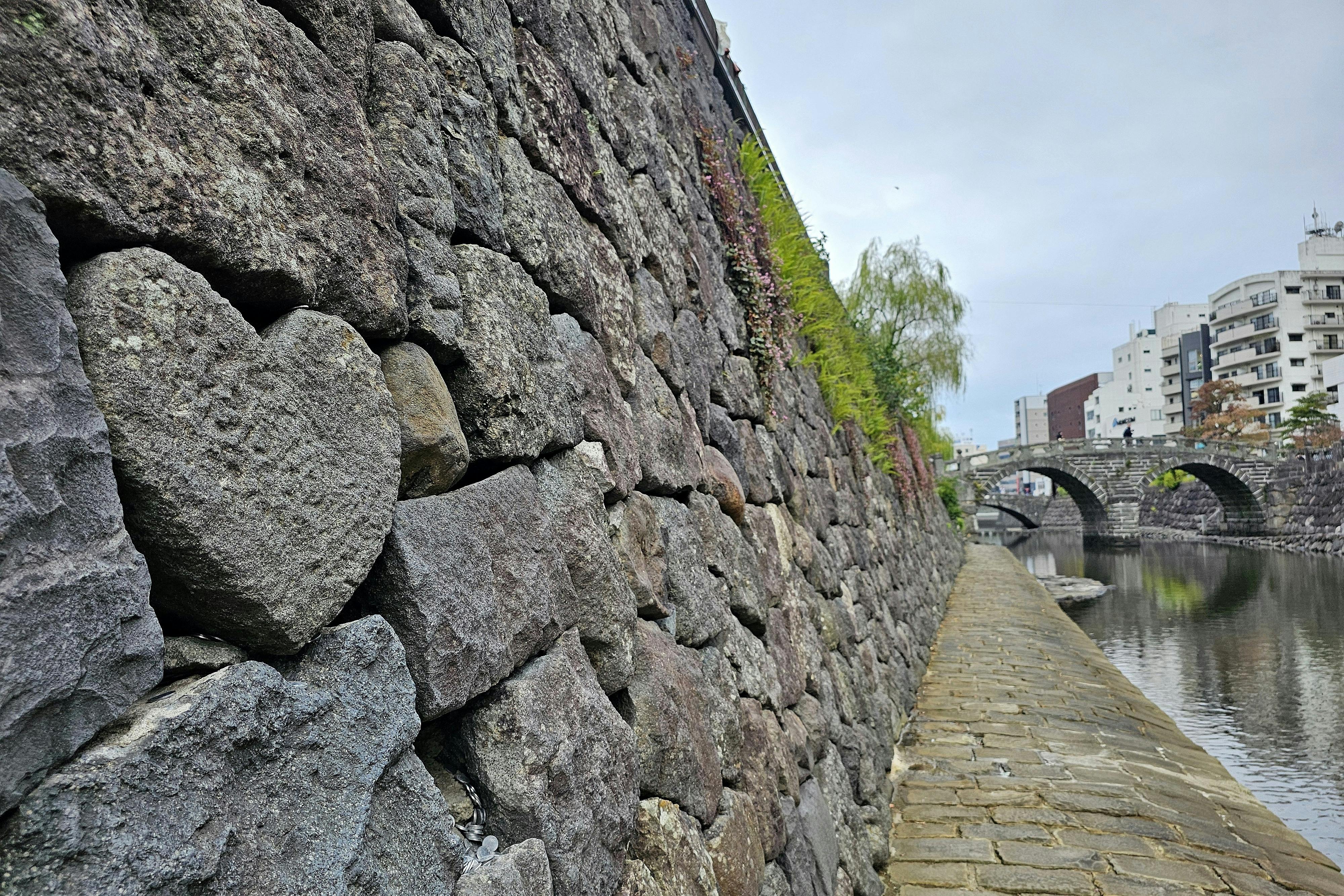 A stone wall along a canal features a heart-shaped rock among the stones, with a walkway beside the water and an arched bridge in the background under an overcast sky.