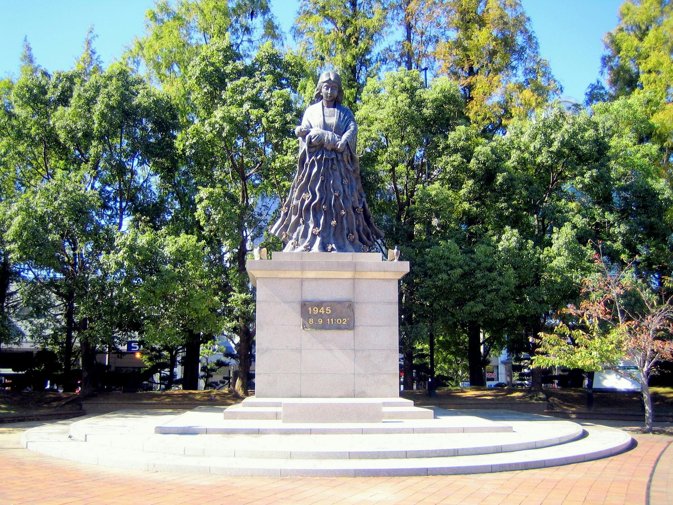 A bronze statue of a person in traditional clothing stands on a stone pedestal with the dates "1945" and "8.9 11:02," surrounded by trees and a sunny blue sky.