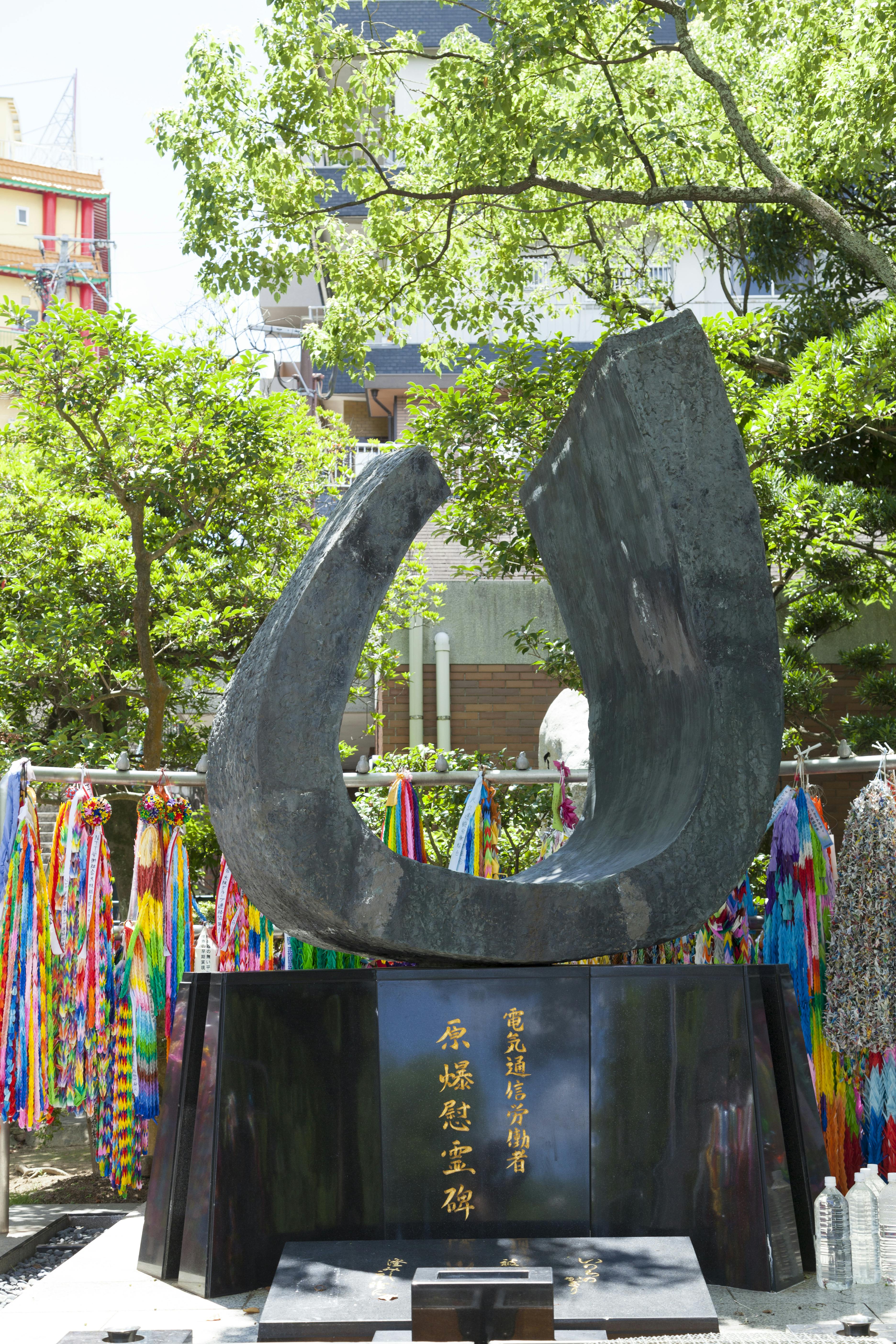 A modern, curved black stone monument with Japanese inscriptions stands on a black pedestal, surrounded by colorful paper cranes and greenery under bright sunlight.