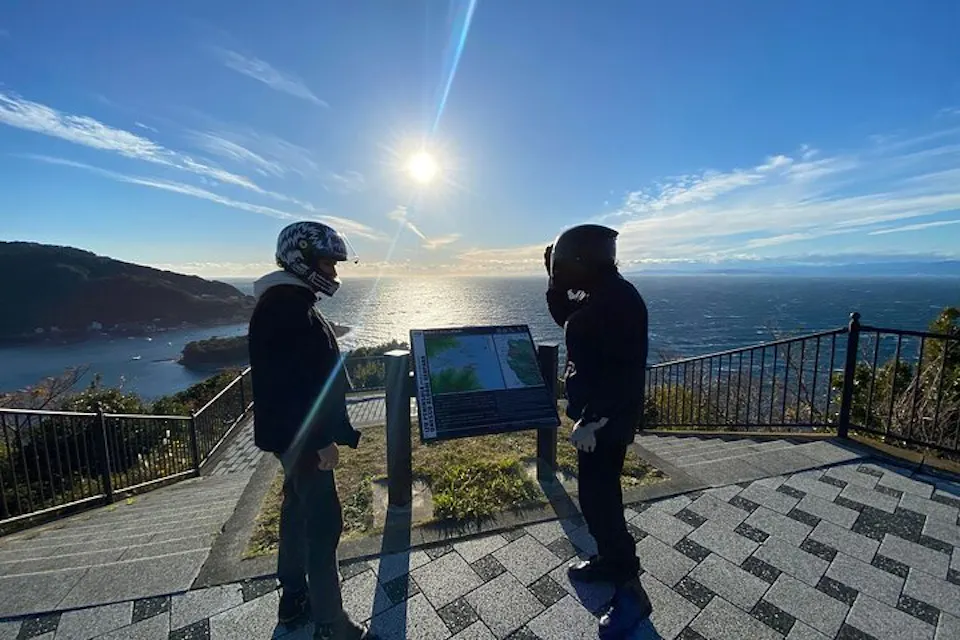 A viewpoint stop on a guided motorcycle day tour in the Hakone area. Two riders in helmets and riding jackets standing at a mountain viewpoint above a lake in bright sunshine