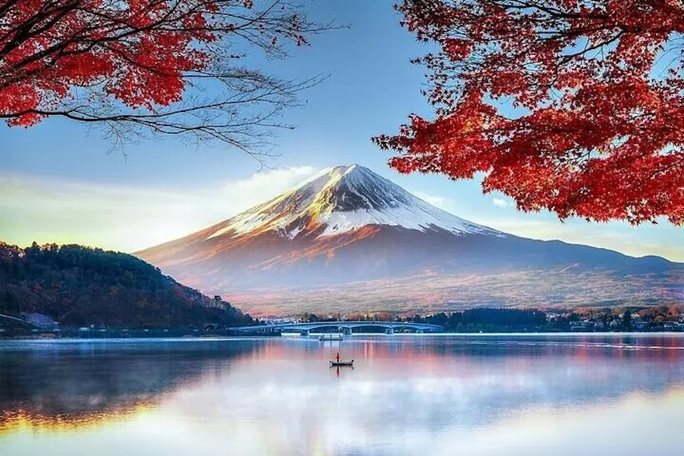 A serene landscape featuring Mount Fuji in the background, partially snow-capped, with vibrant red autumn leaves in the foreground. A calm lake mirrors the scene, with a small boat and two people in the center. A bridge stretches across the lake.