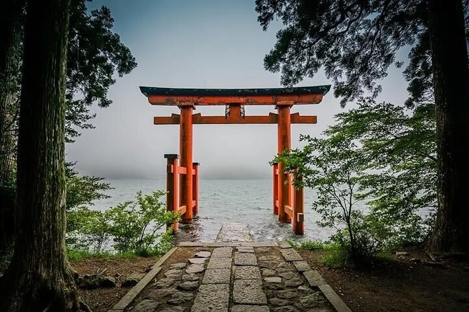 A traditional red torii gate stands at the edge of a serene lake, framed by lush green trees. The pathway leading to the gate is composed of stone slabs, and a misty sky completes the tranquil scene.