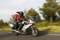 A rider and passenger in full gear on a white adventure motorcycle leaning into a curve on a tree-lined mountain road