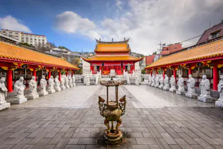 A traditional Chinese temple courtyard with a large incense burner in the center, flanked by rows of white statues under red and gold-roofed walkways, and the main temple building in the background under a cloudy sky.