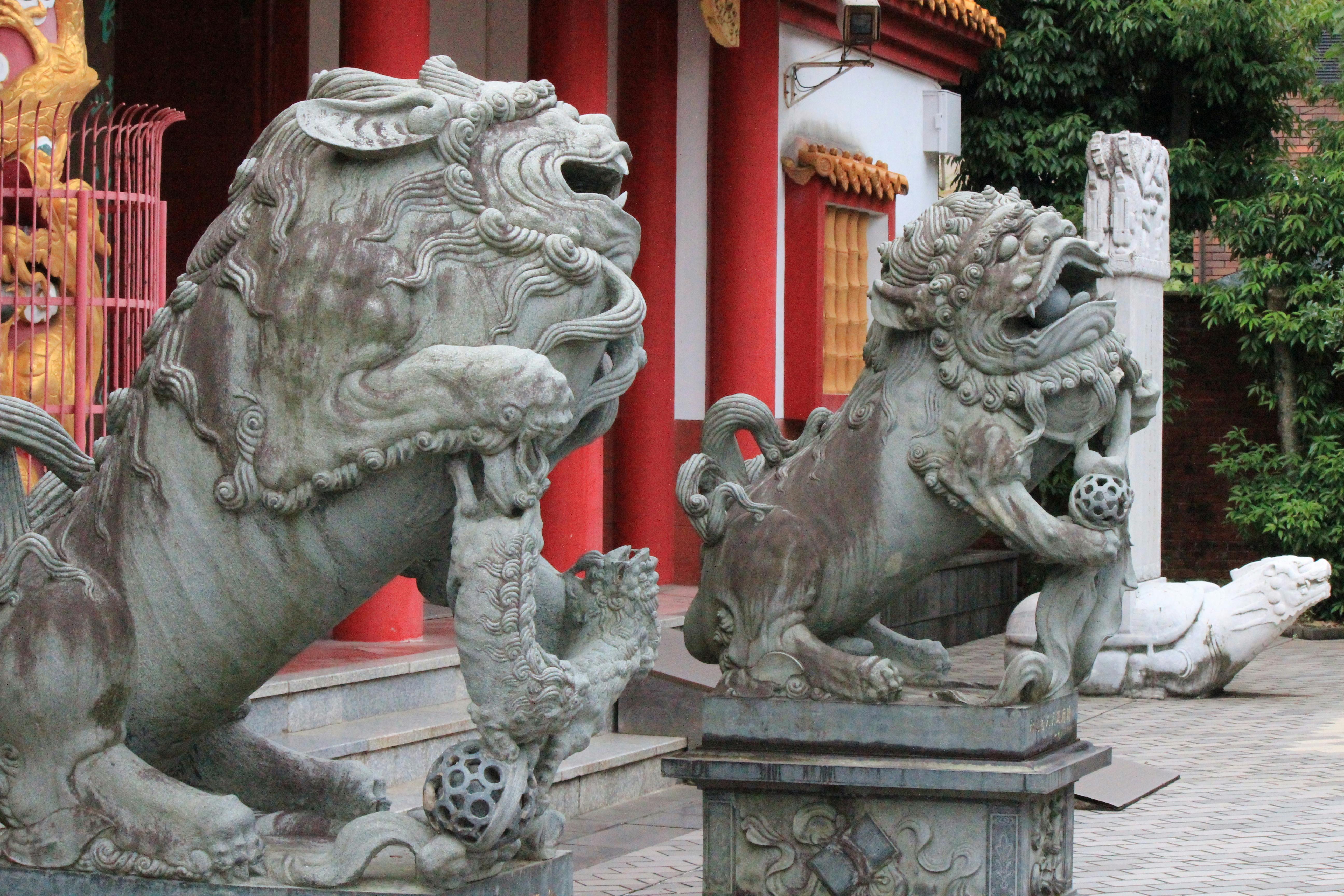 Two stone Chinese guardian lion statues stand in front of a temple with red columns and ornate decorations. One lion has its paw on a ball; the background features trees and another sculpture.