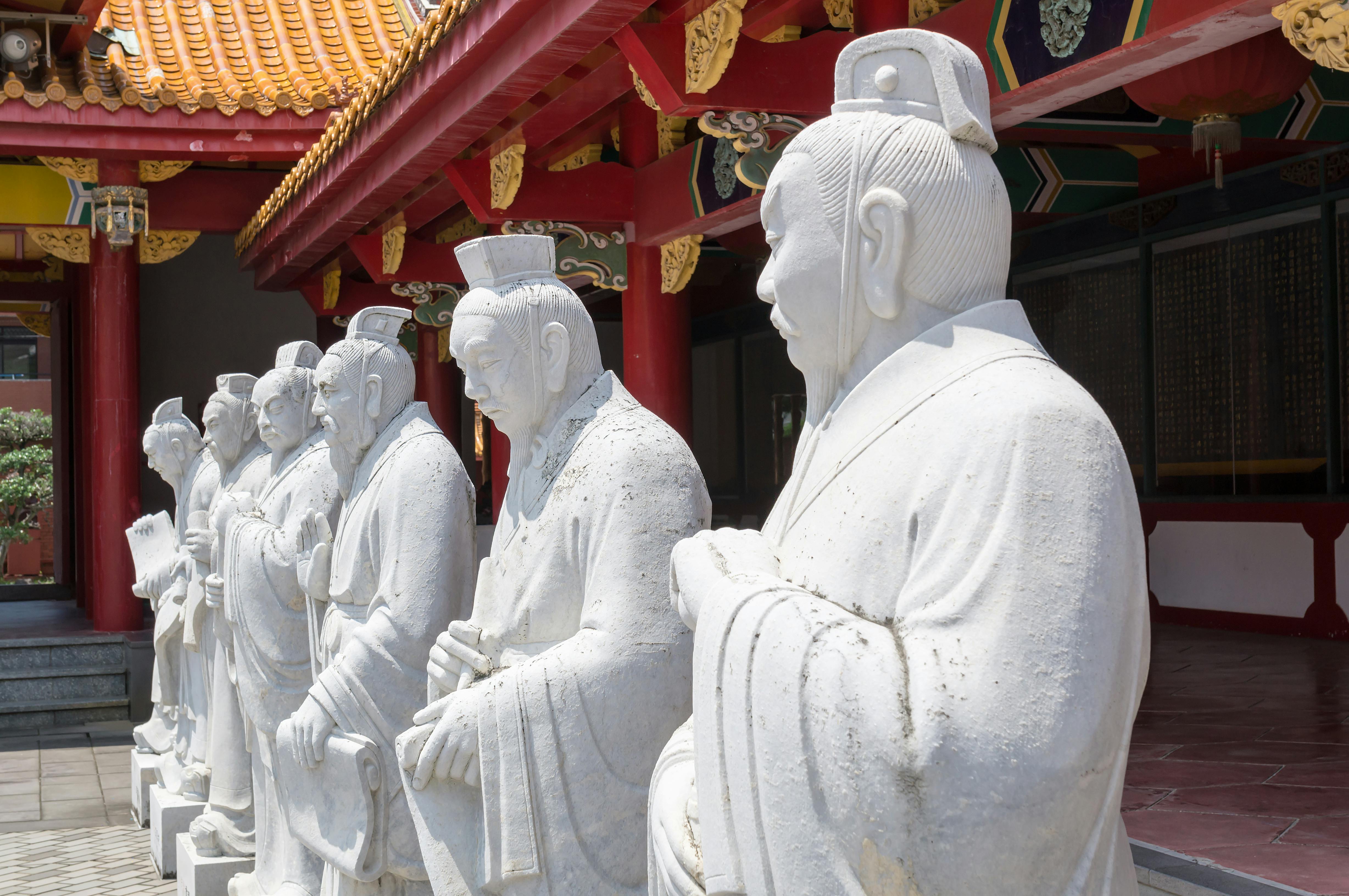 A row of stone statues of ancient Chinese figures stands outside a traditional building with red pillars and a yellow tiled roof.