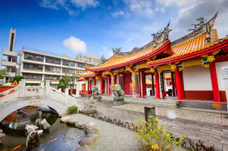 A traditional Chinese temple with ornate red and gold decorations, dragon statues, and a curved stone bridge over a pond, set against a backdrop of modern city buildings under a partly cloudy sky.