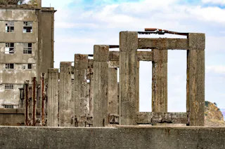Weathered concrete pillars in a row, some rusted with metal supports exposed, stand in front of an old, abandoned building with broken windows. The sky above is partly cloudy.
