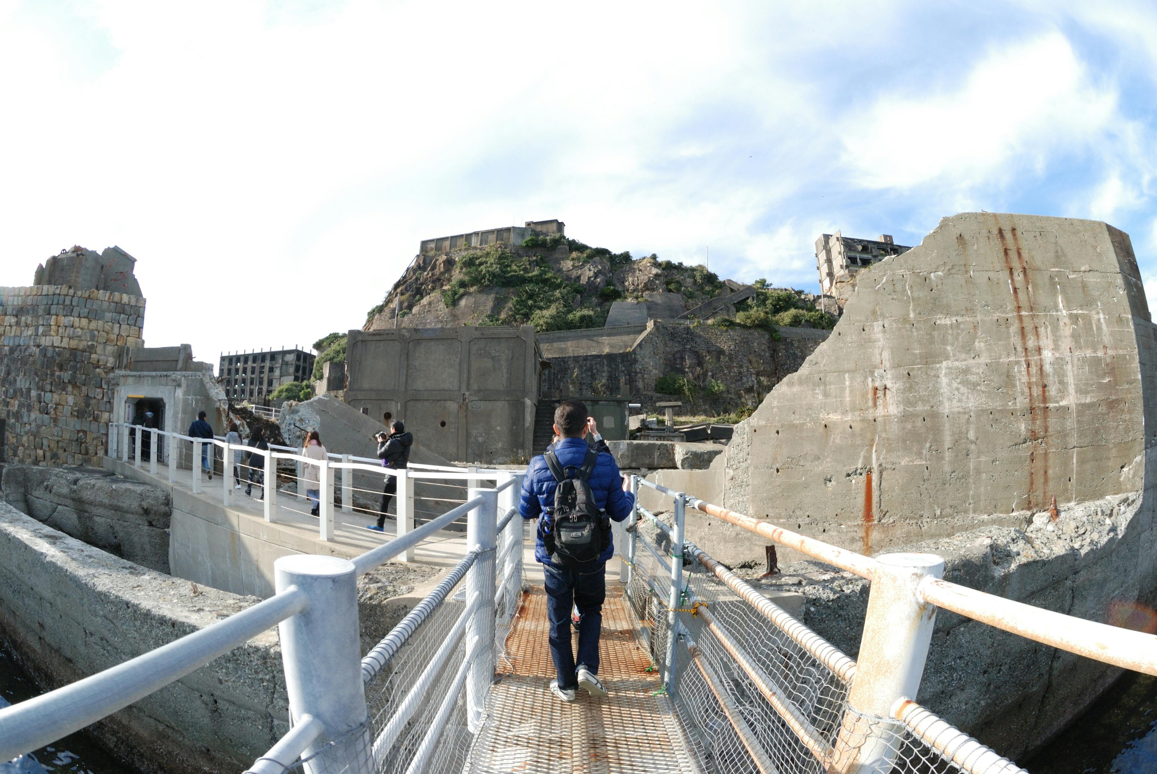 A person with a backpack walks on a metal walkway surrounded by large, weathered concrete structures and ruins, with a few other people in the background and a partly cloudy sky overhead.