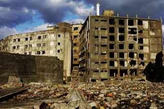 Two large, abandoned concrete buildings with broken windows stand amid rubble and debris under a cloudy sky. The structures appear heavily damaged and weathered, suggesting a neglected or post-apocalyptic scene.
