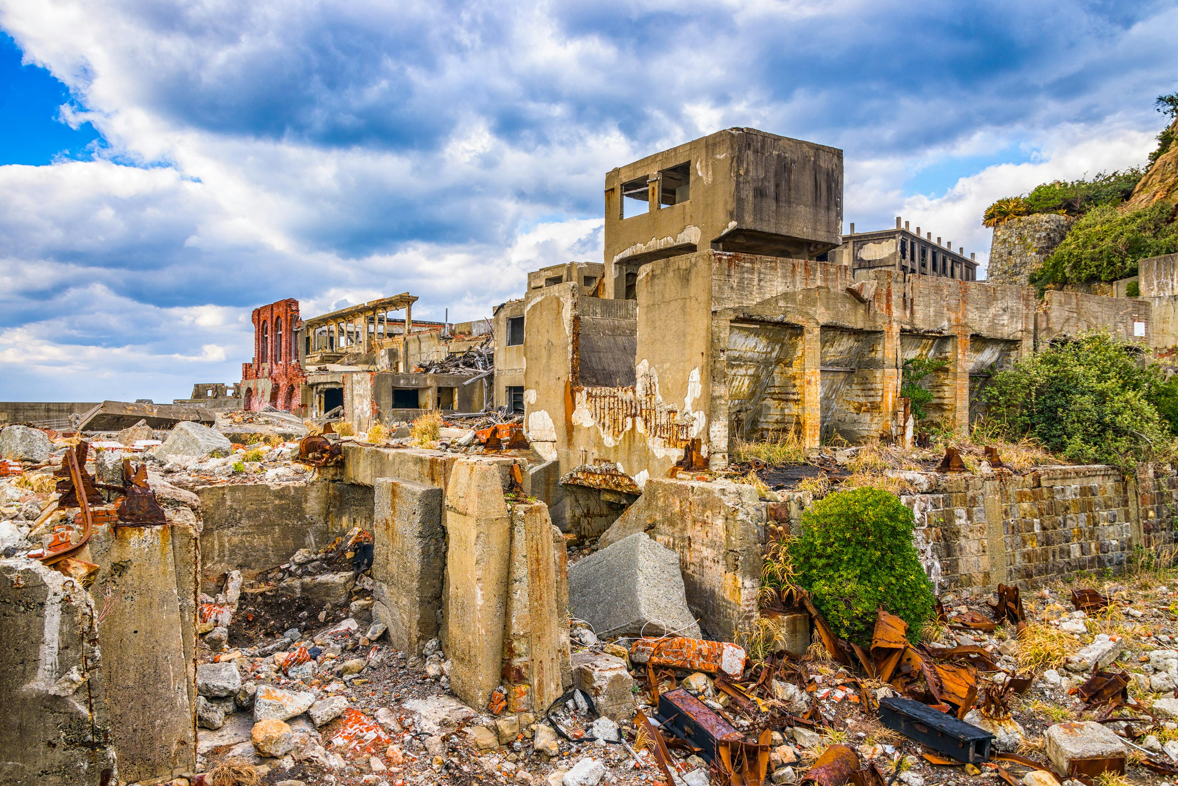 Crumbling concrete buildings and rubble fill an abandoned industrial site under a partly cloudy sky, with overgrown plants and rusted metal debris scattered among the ruins.