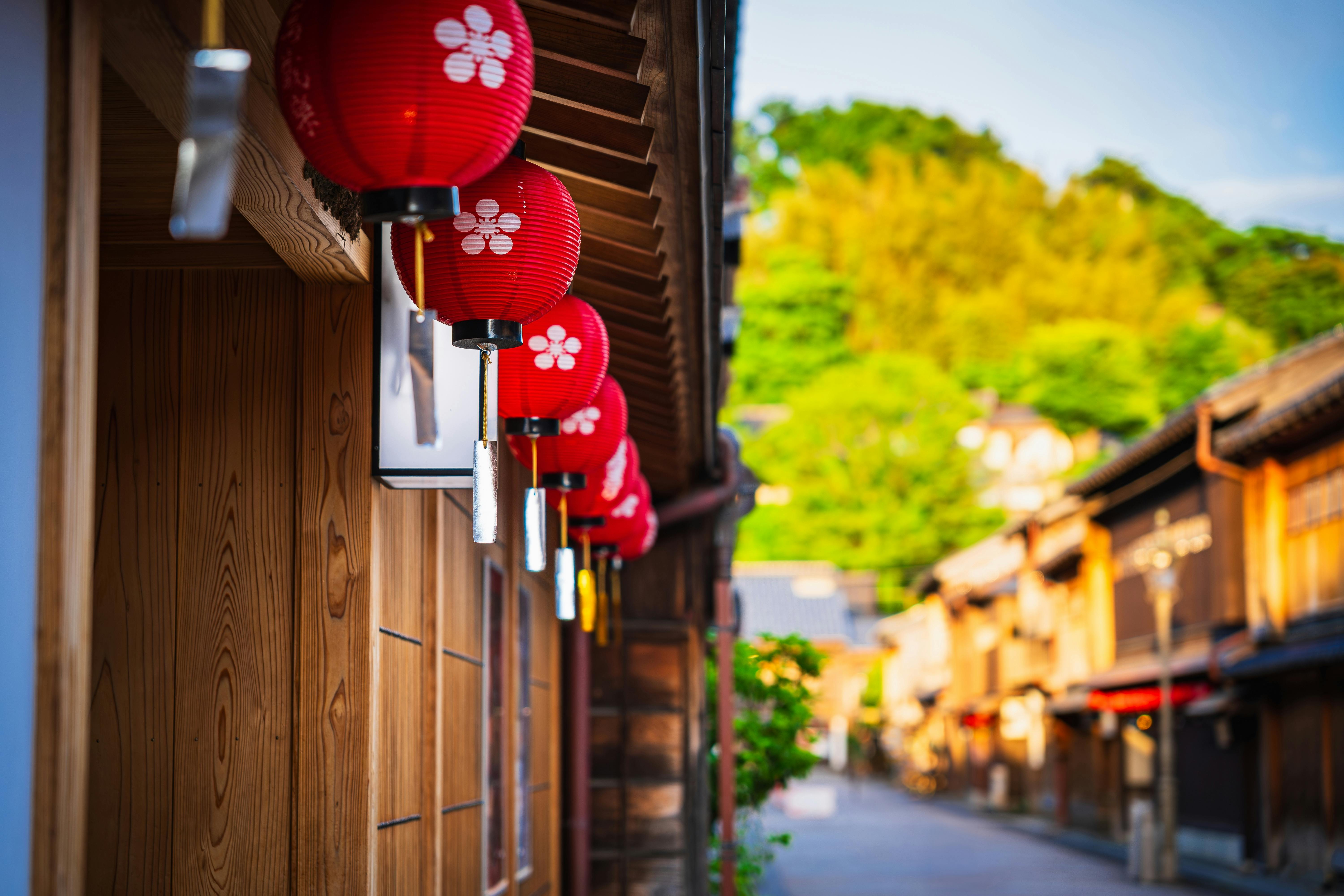 Red paper lanterns with white flower designs hang outside a wooden building along a quiet, sunlit street with traditional Japanese architecture and lush green trees in the background.