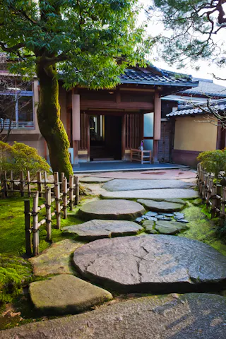 A stone pathway leads through a lush, moss-covered garden, flanked by bamboo fencing, toward a traditional Japanese wooden house with an open entrance and a large tree nearby.
