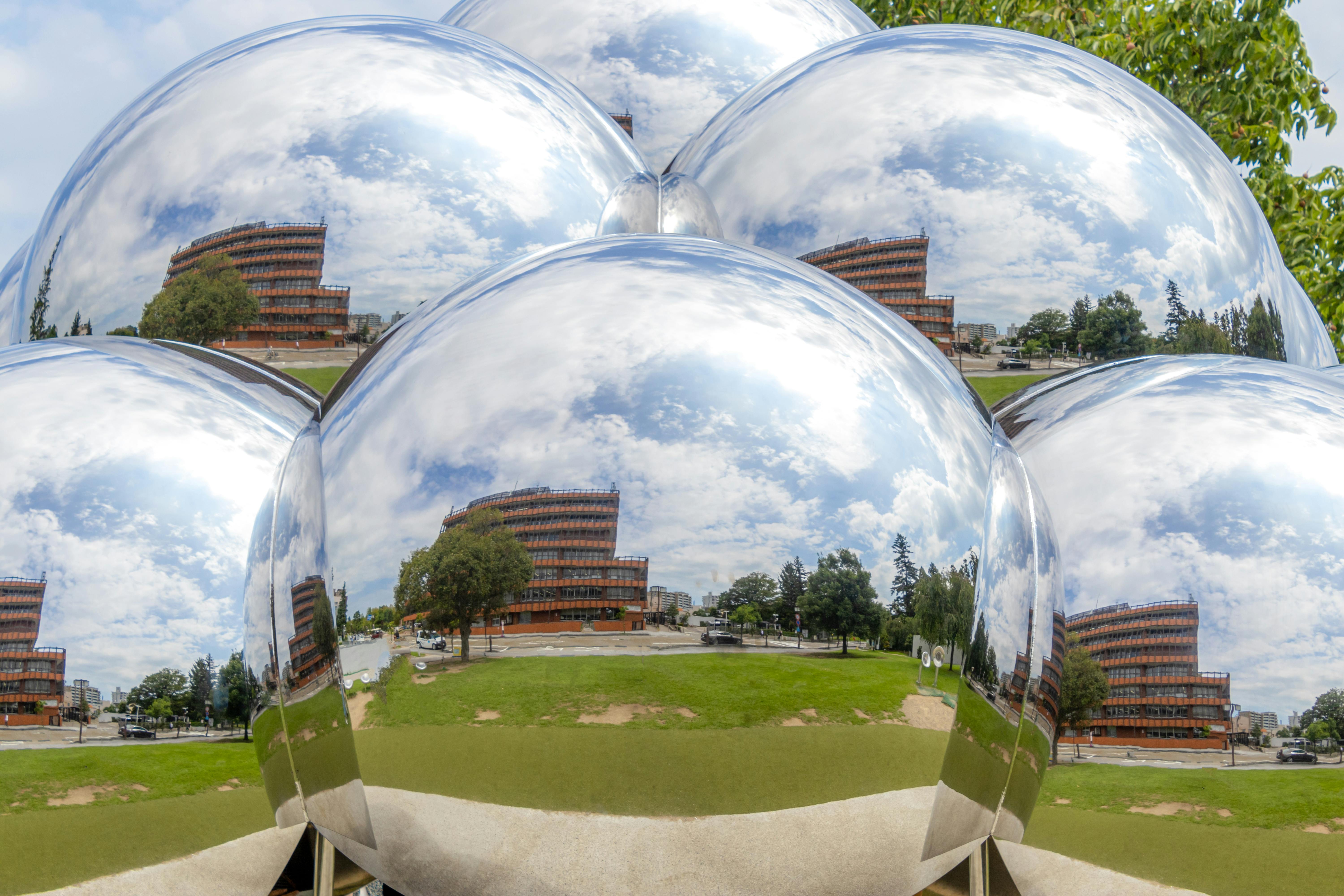 Several large, shiny, silver spheres reflect a red-brick building, green grass, trees, and blue sky with clouds, creating a distorted, artistic mirror effect of the surrounding landscape.