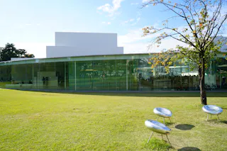 A modern glass building sits behind a grassy lawn with three round, metallic benches and a small tree with sparse leaves under a clear sky.