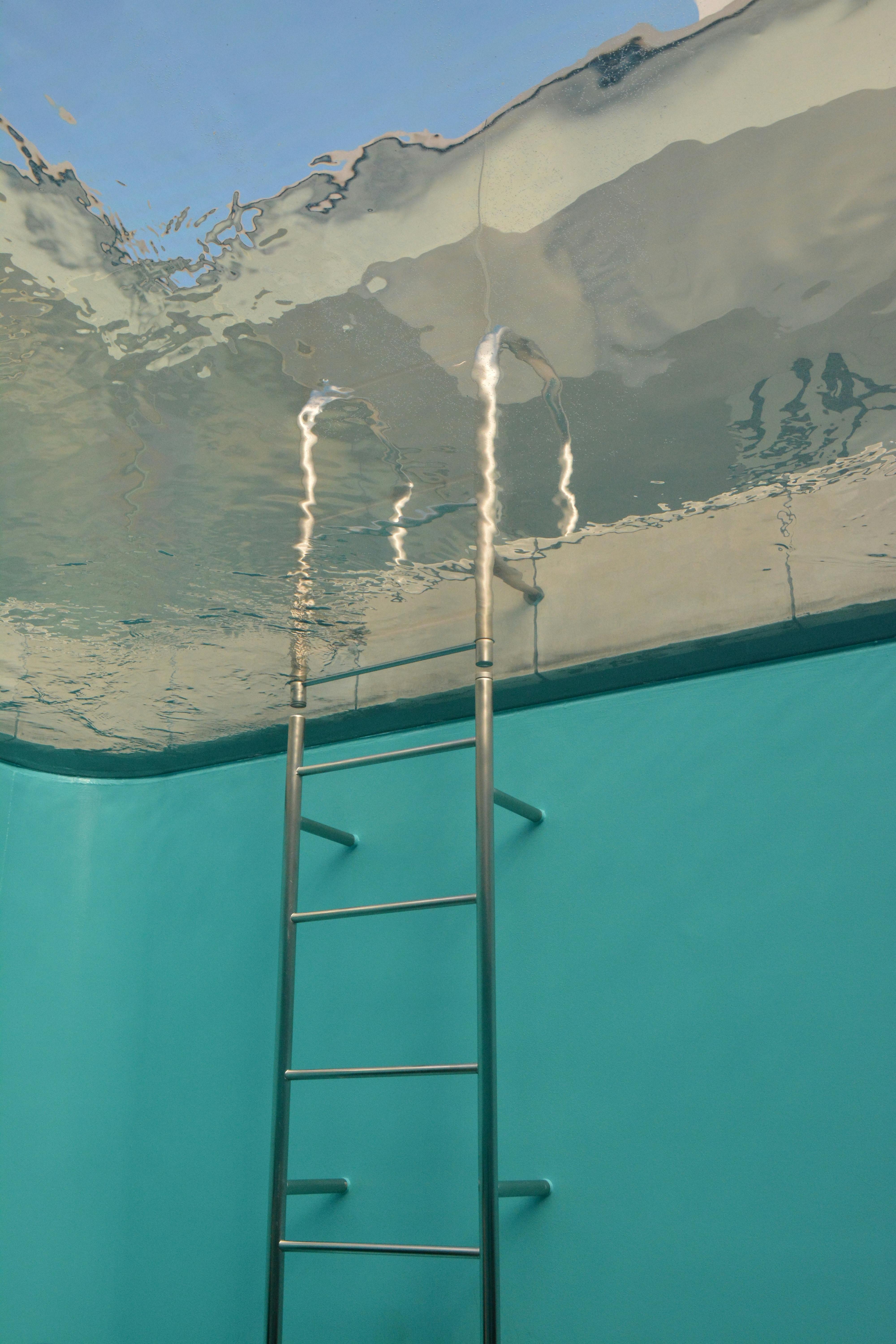 A metal swimming pool ladder leads up to a surface that looks like water, viewed from below. The water appears still and reflective, with light blue pool walls surrounding the ladder.