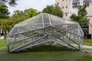 A large geometric metal sculpture with angular, mesh-like panels stands on a grassy area near trees and a multi-story building. The structure resembles a faceted dome or shell.