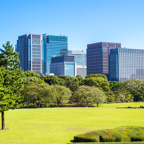 Imperial Palace East Gardens A city skyline with modern high-rise buildings contrasts against a lush, green park in the foreground. Clear blue sky and several trees in the park enhance the vibrant scene on a sunny day.