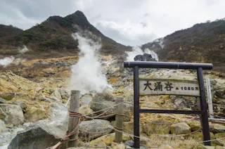 A rocky, steaming volcanic landscape with a wooden sign in Japanese characters and a rope fence in the foreground, set against misty mountains and cloudy skies.