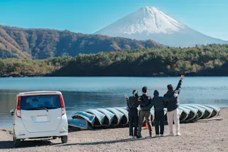 Four people stand by a row of parked boats, facing a scenic view of a lake with a snow-capped mountain in the background. A white car is parked nearby. The sky is clear and blue.