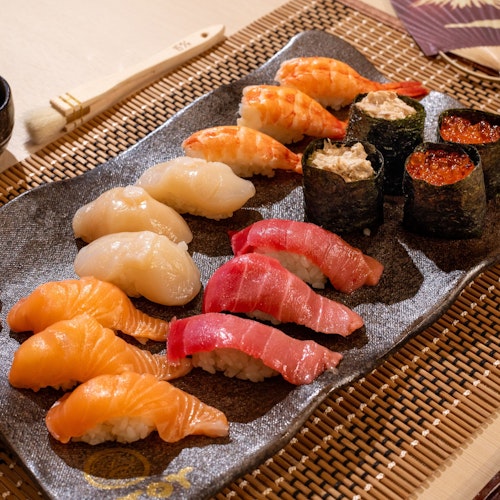 Sushi Making Experience A plate of assorted sushi on a bamboo placemat, featuring salmon, tuna, scallops, shrimp, and roe-topped rolls. Served with pickled ginger, soy sauce in a small cup, and a pair of chopsticks. An open fan is placed beside the plate.